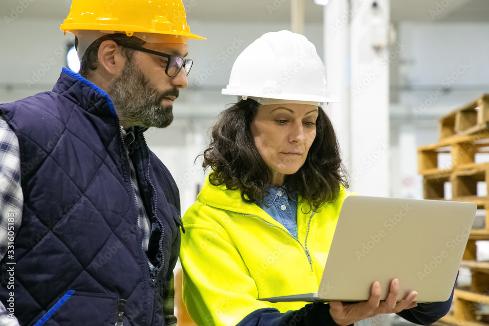 Two factory workers looking at laptop at plant. Confident technicians standing with modern device. Manufacturing concept Two factory workers looking at laptop at plant. Confident technicians standing with modern device. Manufacturing concept
