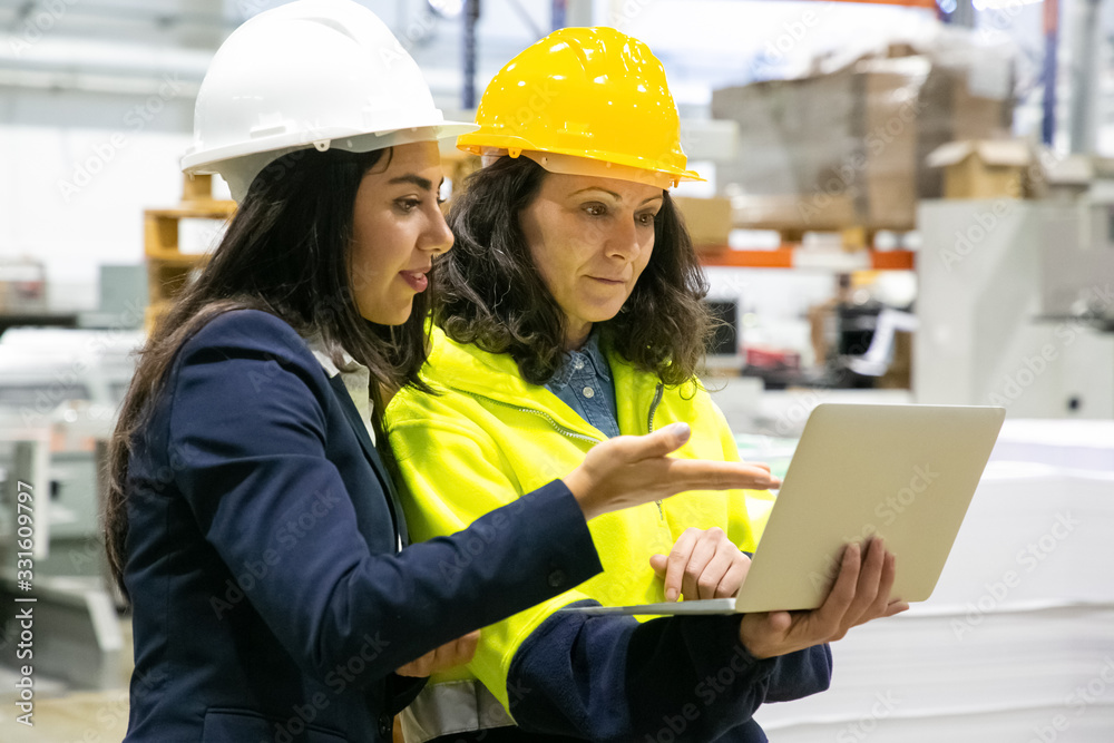 Confident female workers with laptop at manufacturing plant. Two female employees talking while standing at factory with laptop. Print manufacturing concept