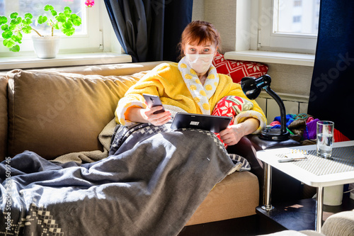 Photo of an adult woman with a viral infection, lying at home on a sofa in a medical mask. Uses a tablet computer