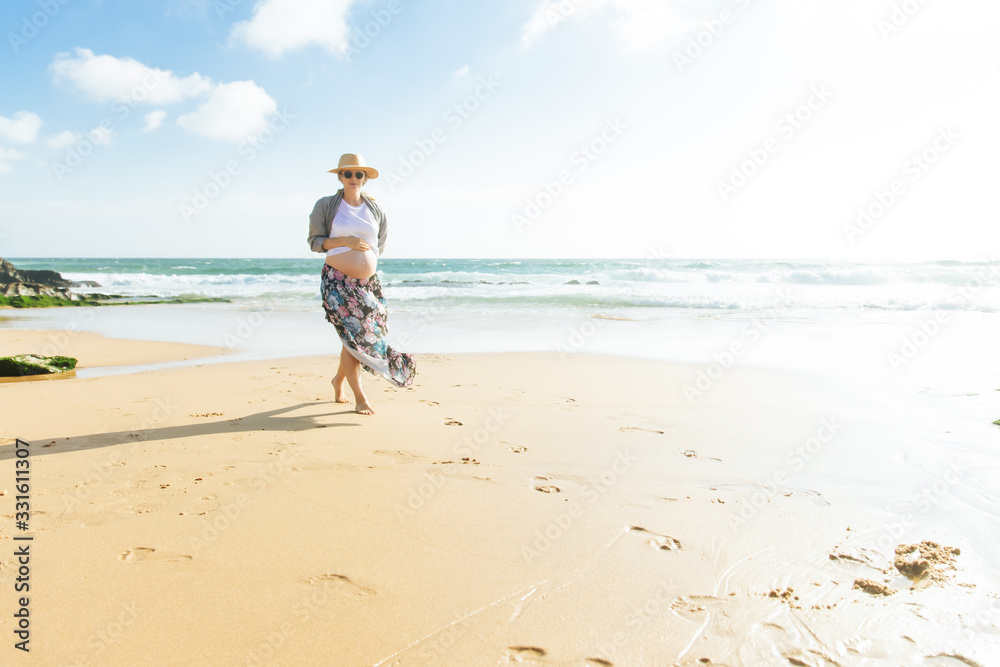 Happy pregnant woman holding hand on belly. Smiling future mother walking on beach during vacation. Pregnancy concept