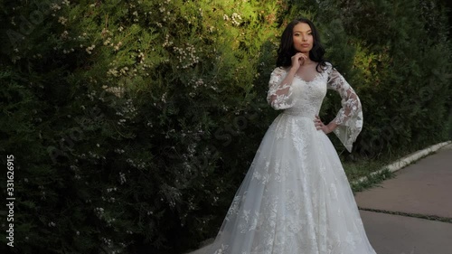 elegant African american bride with loose flowing hair in long lacy wedding dress near high bushes in green park slow motion photo