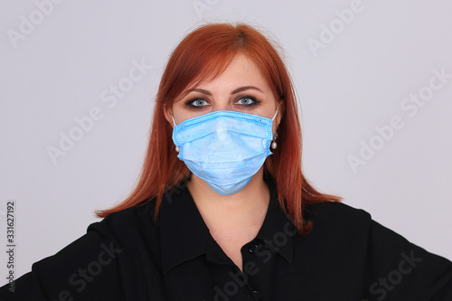 Portrait of a red-haired girl in black in a blue medical mask on a gray background