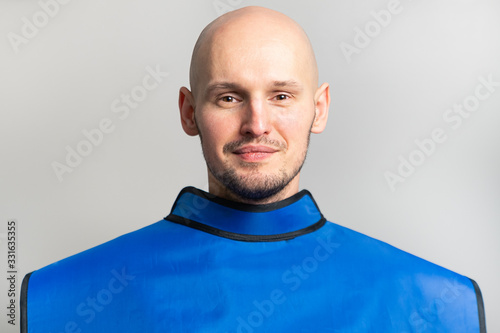 Smiling young bald man with a shaved head in a blue lead apron, close-up portrait on a gray background. X-ray room
