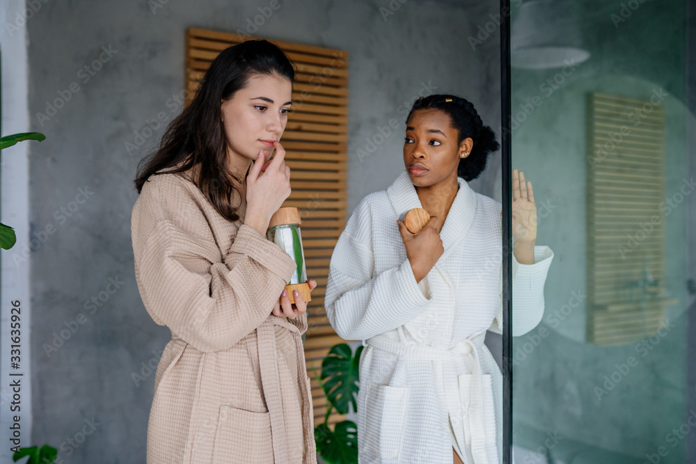 Two multiethnic female friends in bathrobes s relaxing,holding glass jars. Two multiethnic female friends in bathrobes s relaxing,holding glass jars.