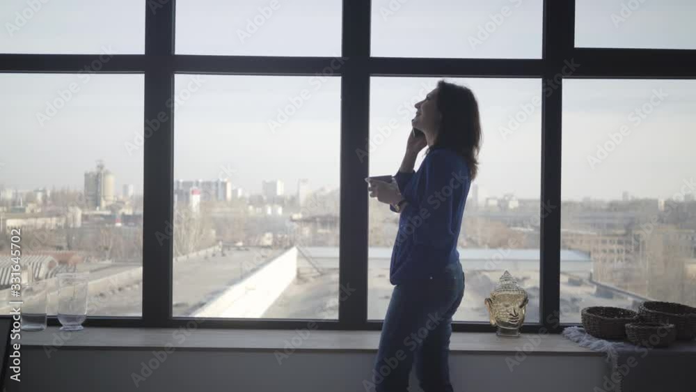 Side view of brunette Caucasian woman with coffee cup standing in front of big window talking on the phone and smiling. Joyful adult businesswoman spending morning indoors. Lifestyle. Side view of brunette Caucasian woman with coffee cup standing in front of big window talking on the phone and smiling. Joyful adult businesswoman spending morning indoors. Lifestyle.