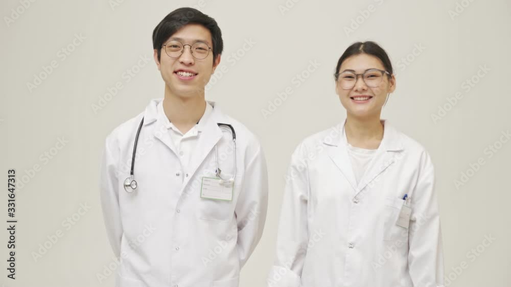 Positive cheery young asian doctors posing isolated over white wall background in sterile uniform showing thumbs up gesture Positive cheery young asian doctors posing isolated over white wall background in sterile uniform showing thumbs up gesture