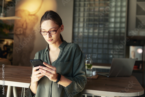 beautiful woman in stylish glasses holding coffee paper cup and looking at smartphone while sitting at cafeteria. Happy woman using mobile phone. Businesswoman drinking coffee and smiling.