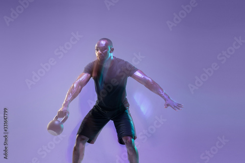 Move. Young african-american bodybuilder training over purple background in neon, mixed light. Muscular male model with the weight. Concept of sport, bodybuilding, healthy lifestyle, motion and action