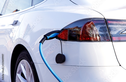 White EV Car or Electric Vehicle at Charging Station with the Power Cable supply plugged in. Eco-Friendly Alternative Green Energy Concept.
