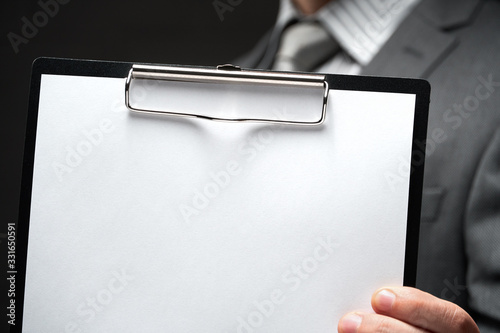 white blank paper sheet closeup in businessman hand, gray suit, dark wall background, blank space for text