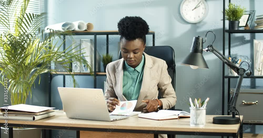 African American beautiful busy female worker looking through graphs and typing on smartphone at office. Occupied pretty smart employee studying documents at workplace and tapping on mobile phone African American beautiful busy female worker looking through graphs and typing on smartphone at office. Occupied pretty smart employee studying documents at workplace and tapping on mobile phone