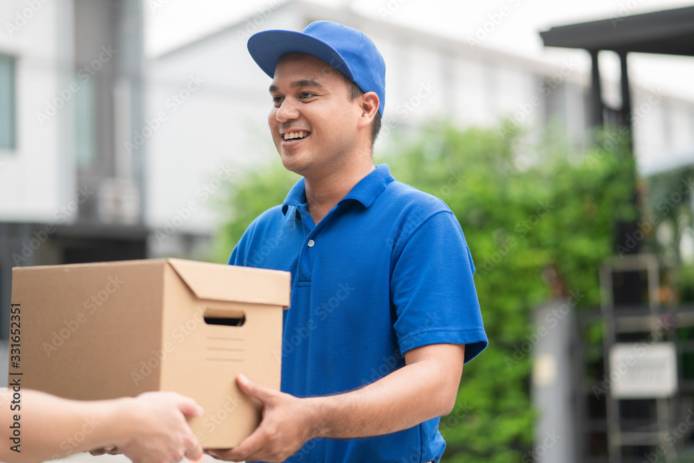 A smiling young asian delivery in blue uniform with parcel cardboard in front of customer house. Messenger and delivery concept. A smiling young asian delivery in blue uniform with parcel cardboard in front of customer house. Messenger and delivery concept.