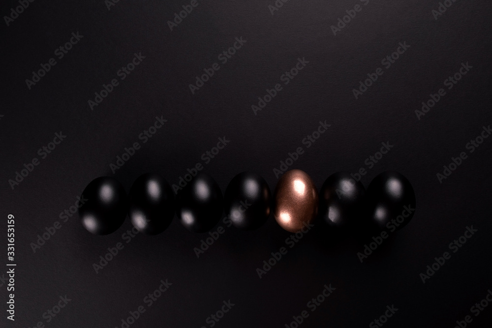 One golden egg and black eggs lie in a row on black background with copy space