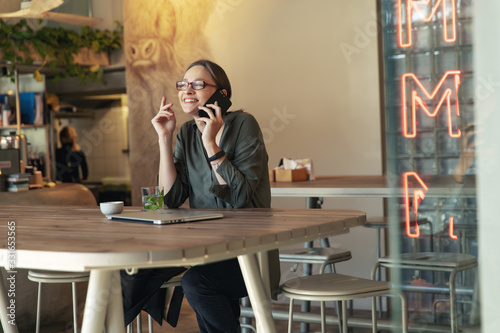 Attractive businesswoman in black jacket and stylish glasses sitting in cafe, talking on the phone and looking at laptop.
