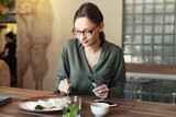 Business woman in stylish glasses sitting with a laptop in a cafe and eating breakfast with a cup of coffee on the table.