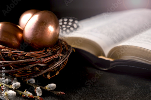 Open Bible on a dark table, eggs in a nest  and feather on dark background. Easter greeting card