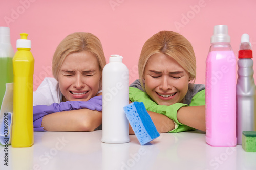 Indoor photo of sad young blonde housewives being exhausted after spring cleaning and frowning their faces while crying with closed eyes, posing over pink background photo