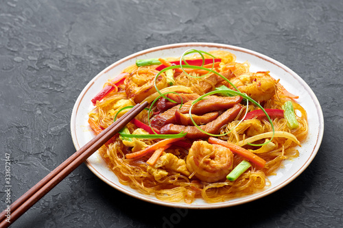 Singapore Mei Fun in white plate on dark slate background. Singapore Noodles is chinese cuisine dish with rice noodles, prawns, char siu pork, carrot, red onion, napa cabbage. Chinese food.