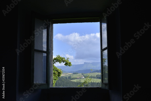 Paisaje verde con bosques y prados y la sierra en el horizonte visto desde el interior de una casa oscura a través de una vieja ventana