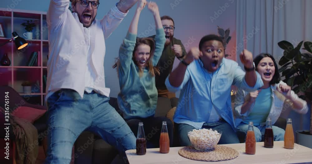 Group of cheerful people sitting on sofa and watching sports on TV at home. Mixed raced youth supporters shouting, applouding and giving high five while celebrating success of their team.