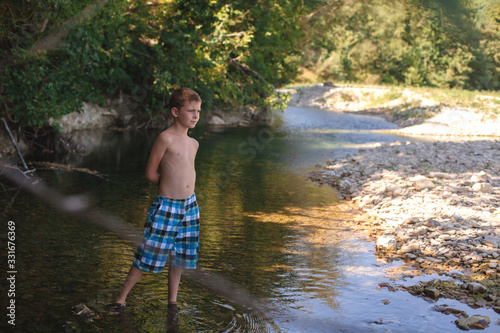 A teenage boy in shorts plays in a mountain river in shallow water