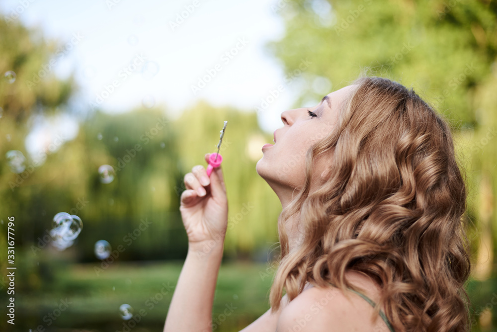 Close-up portrait of young pretty woman with blond curly hair, blowing soap bubbles in green park in summer. Summertime fun. Making colorful soap bubbles. Close-up portrait of young pretty woman with blond curly hair, blowing soap bubbles in green park in summer. Summertime fun. Making colorful soap bubbles.