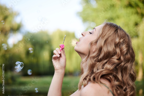 Close-up portrait of young pretty woman with blond curly hair, blowing soap bubbles in green park in summer. Summertime fun. Making colorful soap bubbles.