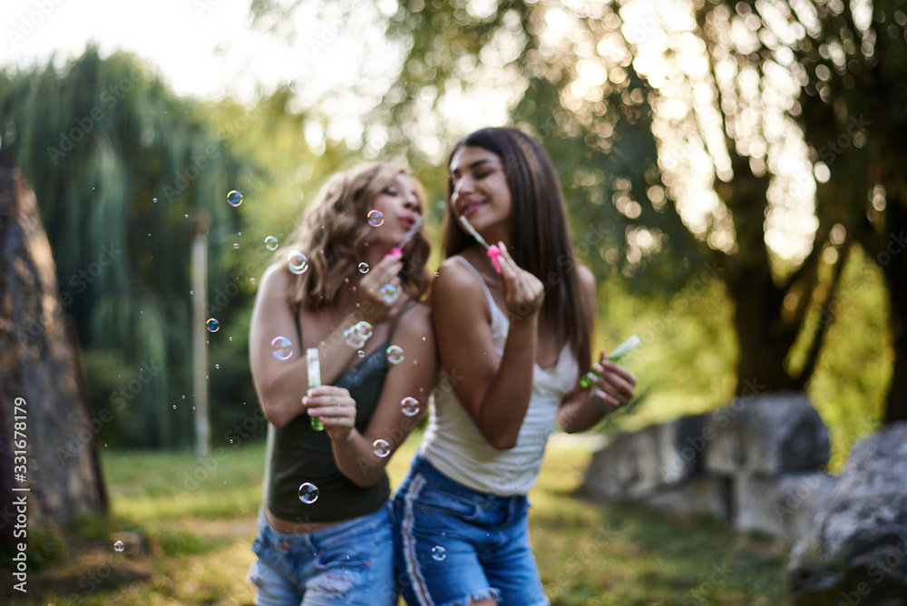 Two sisters, blowing soap bubbles in park, Young pretty girls, wearing jeans shorts, green khaki beige tops, having fun outside. Three-quarter portrait of two girlfriends making soap bubbles, smiling.