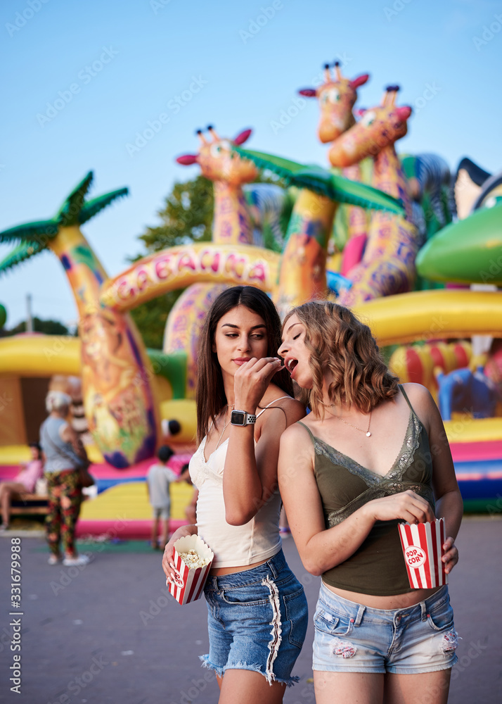 Fototapeta premium Two sisters, standing is front of attraction in theme park, holding popcorn in paper box, smiling, having fun. Pretty girls. wearing blue jeans and beige and green tops, relaxing in amusement park