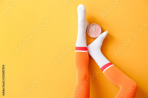 Legs of young woman in socks and with alarm clock on color background photo