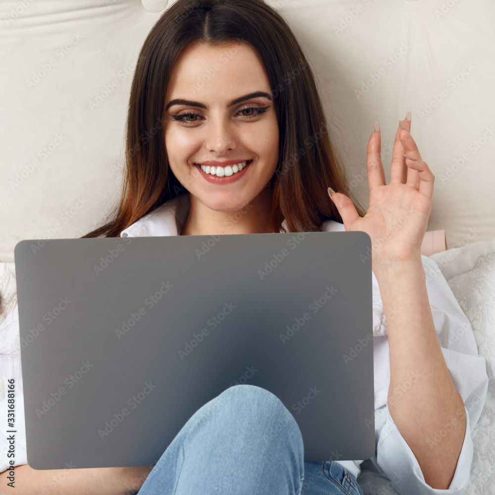 Girl waving hand to video call on laptop laying in bed