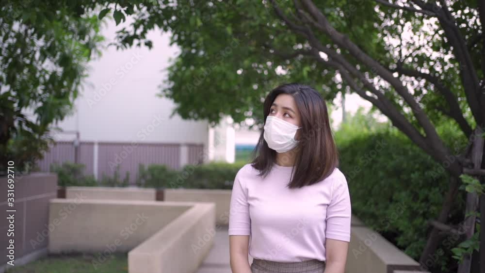 Young adult Asian woman wear white mask walking toward camera at outdoor park walkway under trees shades, alone home quarantine, covid19 corona virus, front view, 1080p slow motion loneliness feeling
