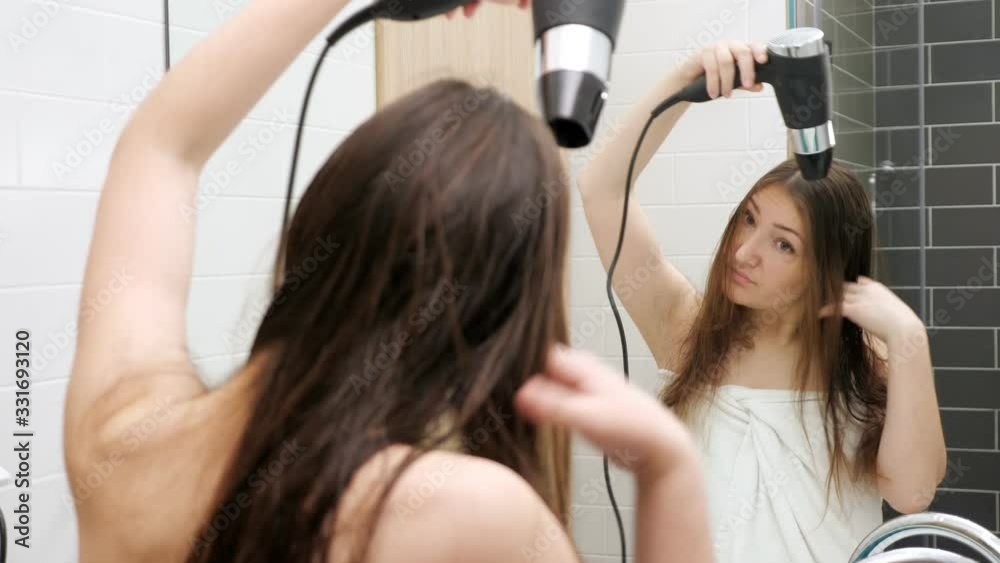 attractive brunette in white towel dries long loose hair with modern black hairdryer looking into mirror close view attractive brunette in white towel dries long loose hair with modern black hairdryer looking into mirror close view