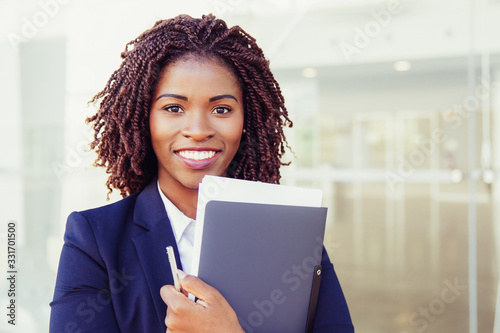 Happy friendly office assistant posing outside. Young black business woman standing at glass wall, holding documents, looking at camera, smiling. Assistant or paperwork concept