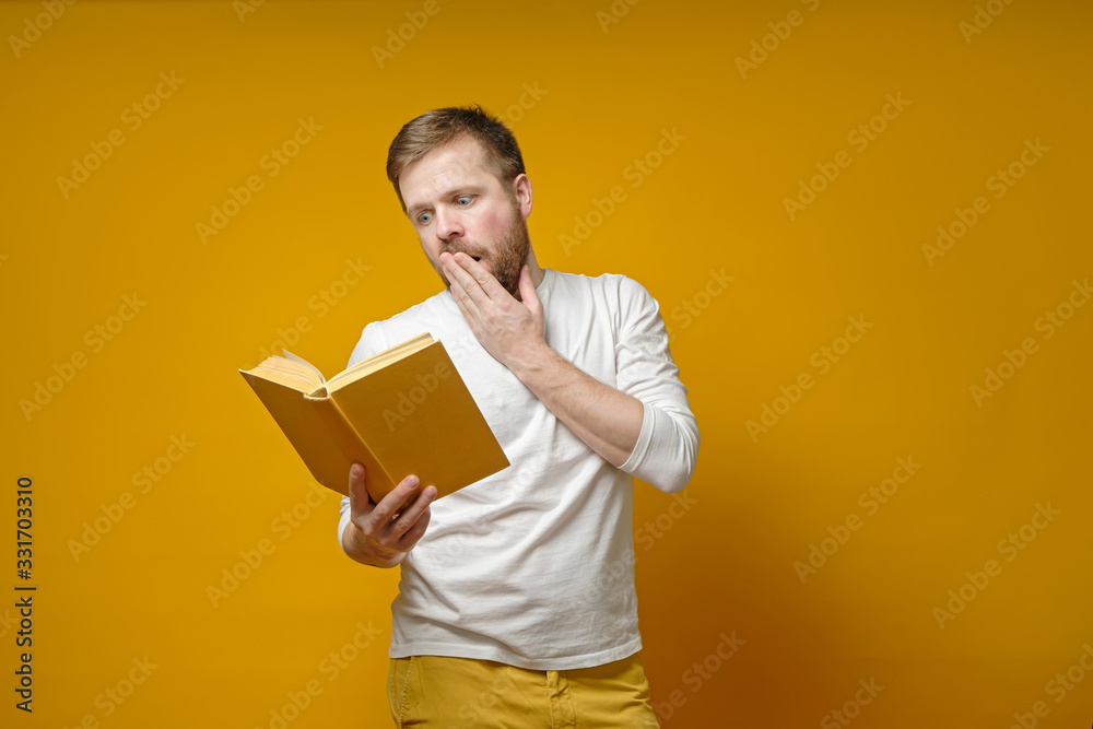 Young, bearded man is fascinated reading a book and emotionally covers his mouth with his hand, on a yellow background.