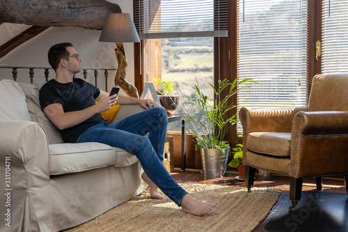 Young man with a cell phone in his hand sitting on a couch in a rustic house looking out the window