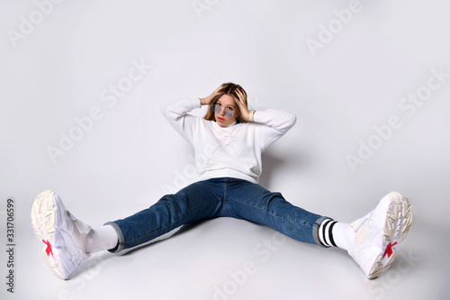 Teenager in sunglasses, jeans, sweater, socks and sneakers. Posing sitting on floor, legs wide apart, isolated on white. Close up