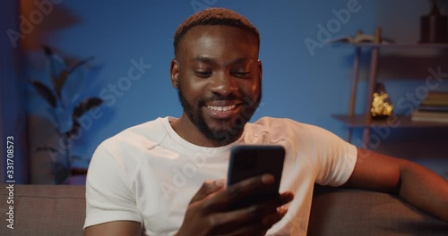 Smiling young man using frontal camera on his smartphone for chatting with friend. Happy guy sitting on sofa, talking and gesturing while having video call. Home background. photo