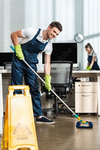 cheerful cleaner washing floor with mop while smiling at camera