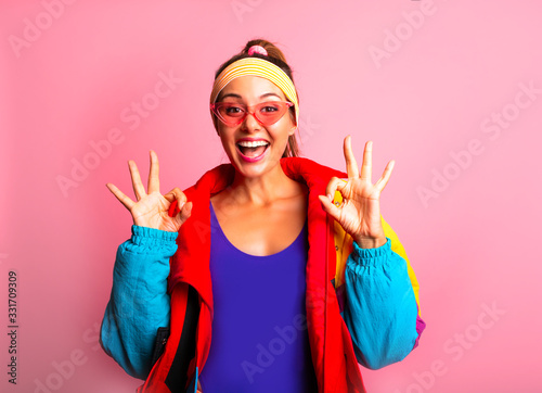 Photo Portrait of happy woman in colourful sport coat, violet fitness body suit and sunglasses of 80s 90s on pink background showing Ok