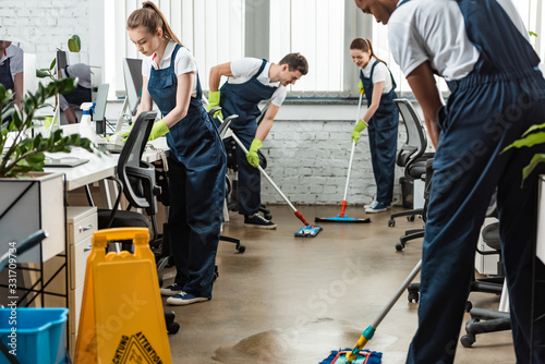 multicultural team of young cleaners washing floor with mops in office