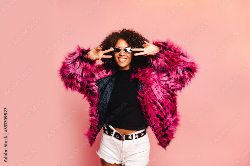 Studio shot of joyful Afro American woman laughs sincerely, feels relaxed, dances while listens favourite music, wears pink fur coat and sparkling dress, models over rosy background. Celebration