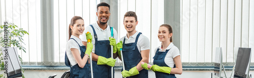 panoramic shot of cheerful multicultural team of cleaners looking at camera while standing in office photo