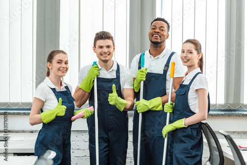 happy multicultural team of cleaners looking at camera and showing thumbs up photo