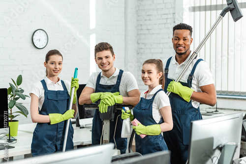 Fotografía happy multicultural cleaners looking at camera while standing with cleaning supp
