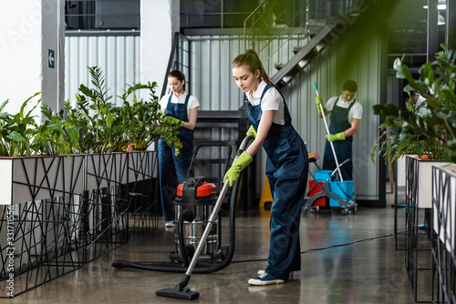 Фотография young cleaner vacuuming floor in office near colleagues