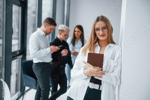 Portrait of cheerful blonde with notepad that stands in front of young successful team that working and communicating together indoors in office