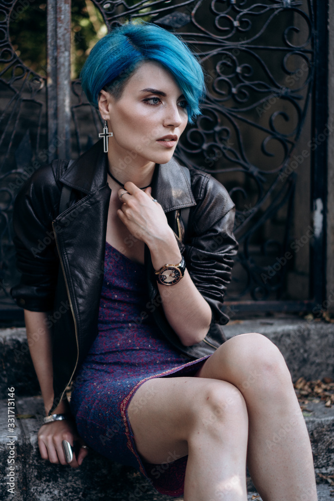 Fototapeta premium Portrait of a girl in a dress and a black leather jacket sitting on the steps near the forged gate