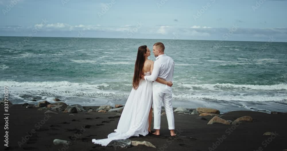 Beautiful beachside with a black sand standing in front of the seaside amazing couple in love they have a romantic time kissing each other with passion Beautiful beachside with a black sand standing in front of the seaside amazing couple in love they have a romantic time kissing each other with passion