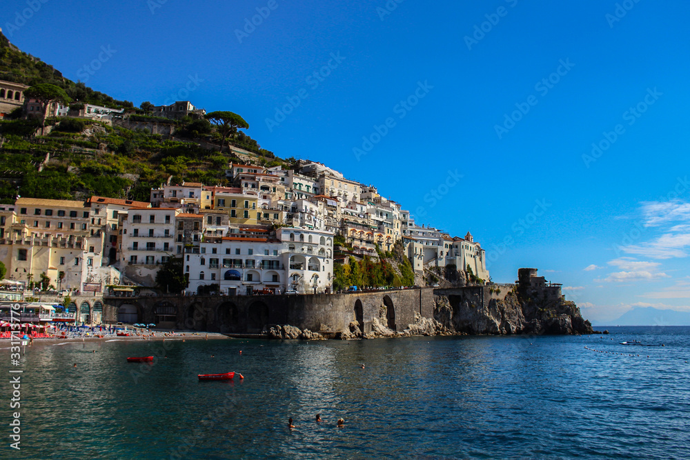 View of the beach with its bathers from the town of Amalfi from the jetty with the sea, boats and colorful houses on the slopes of the Amalfi coast in the province of Salerno, Campania, Italy.